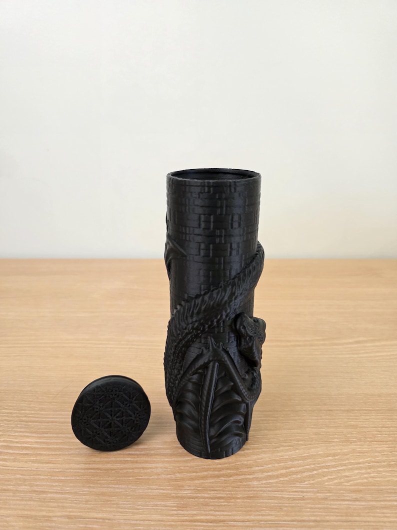May include: A black cylindrical vase with a detailed dragon design wrapped around it. The vase has a matching lid with a geometric pattern. The vase is approximately 6 inches tall.