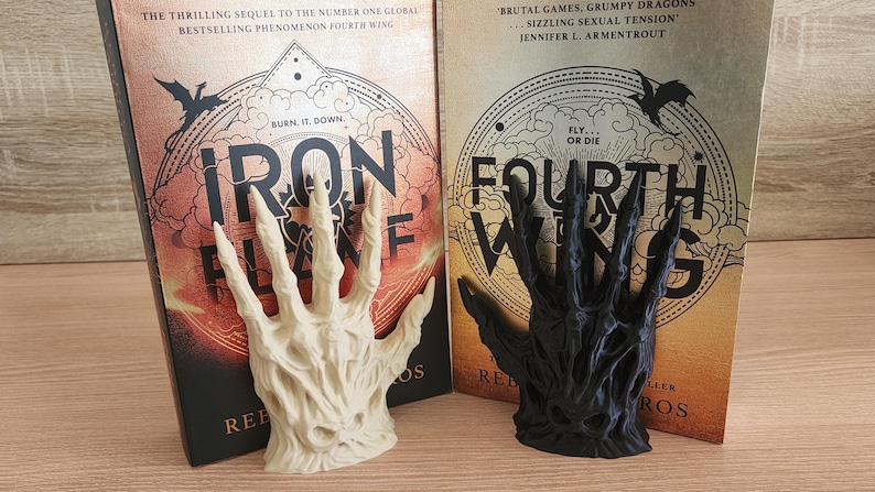 Peut inclure: Deux serre-livres en forme de mains squelettiques, l'un blanc cass&eacute; et l'autre noir, devant deux livres. Les livres sont intitul&eacute;s "Iron Flame" et "Fourth Wing". Les couvertures pr&eacute;sentent des dragons et des fonds flamboyants.