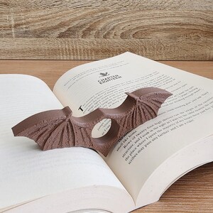 May include: A brown, winged bookmark shaped like a bat, with a textured surface, is placed in a book. The book is open to a page with the text "Chapter Eighteen".