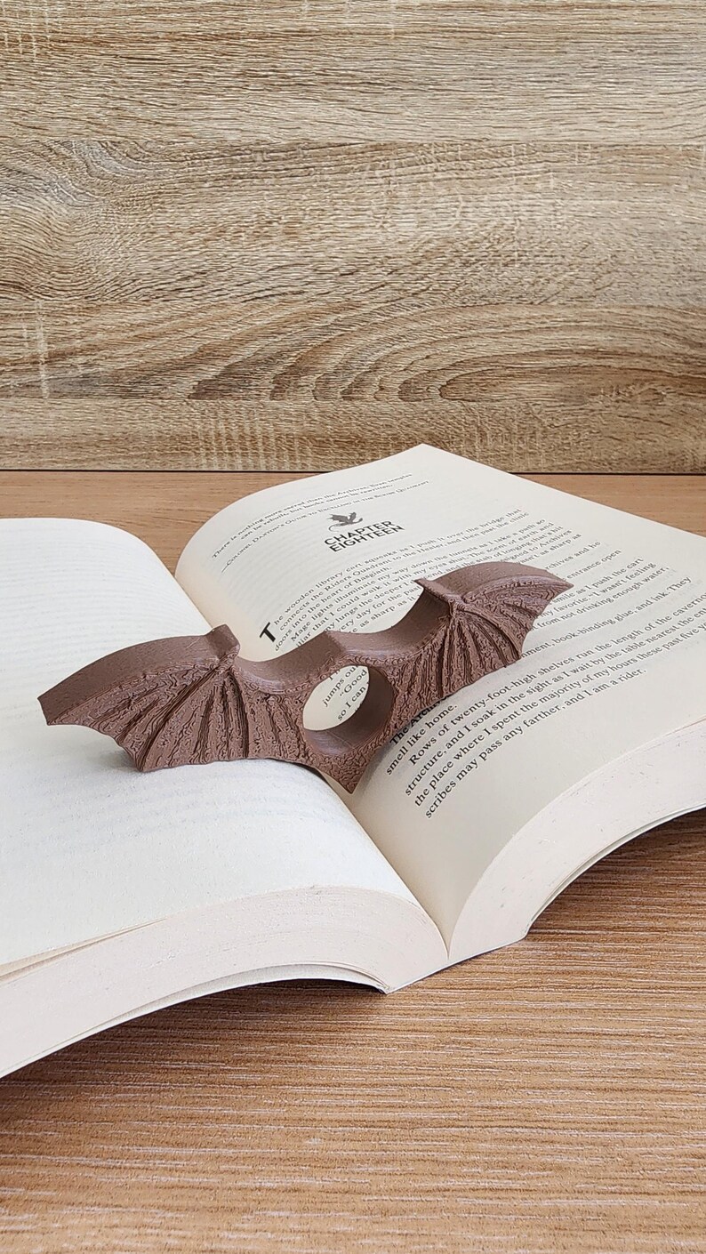 May include: A brown, winged bookmark shaped like a bat, with a textured surface, is placed in a book. The book is open to a page with the text "Chapter Eighteen".
