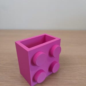 Peut inclure: Un organiseur de bureau rose vif, ressemblant &agrave; une grosse brique de construction. Il comporte quatre tenons circulaires en relief sur un c&ocirc;t&eacute; et est fabriqu&eacute; en plastique solide. L'int&eacute;rieur est creux, offrant de l'espace pour stylos, crayons ou petits objets.