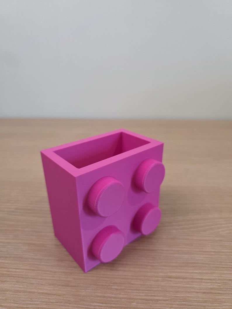 Peut inclure: Un organiseur de bureau rose vif, ressemblant &agrave; une grosse brique de construction. Il comporte quatre tenons circulaires en relief sur un c&ocirc;t&eacute; et est fabriqu&eacute; en plastique solide. L'int&eacute;rieur est creux, offrant de l'espace pour stylos, crayons ou petits objets.