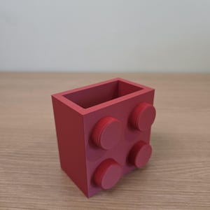 Peut inclure: Un organiseur de bureau rouge en forme de grosse brique de construction. Il comporte quatre tenons circulaires sur un c&ocirc;t&eacute; et un int&eacute;rieur creux. L'organisateur est fait d'un mat&eacute;riau solide. Dimensions d'environ 8 cm x 8 cm x 8 cm.