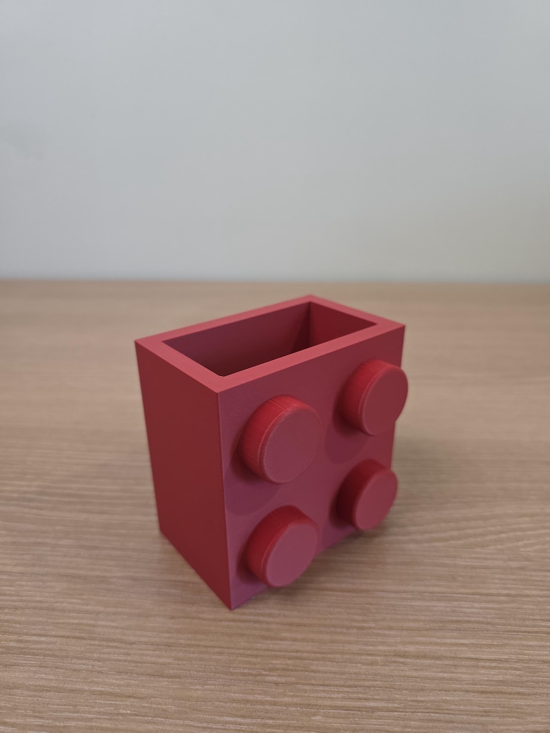 Peut inclure: Un organiseur de bureau rouge en forme de grosse brique de construction. Il comporte quatre tenons circulaires sur un c&ocirc;t&eacute; et un int&eacute;rieur creux. L'organisateur est fait d'un mat&eacute;riau solide. Dimensions d'environ 8 cm x 8 cm x 8 cm.