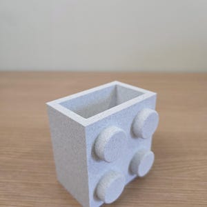 Peut inclure: Un r&eacute;cipient rectangulaire gris clair avec quatre plots circulaires en relief sur un c&ocirc;t&eacute;. Le r&eacute;cipient a une texture mouchet&eacute;e et est pos&eacute; sur une surface en bois. Le design ressemble &agrave; une brique de construction.