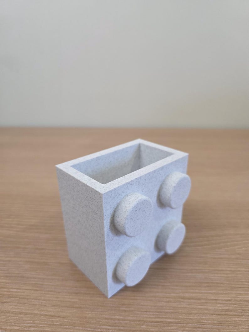 Peut inclure: Un r&eacute;cipient rectangulaire gris clair avec quatre plots circulaires en relief sur un c&ocirc;t&eacute;. Le r&eacute;cipient a une texture mouchet&eacute;e et est pos&eacute; sur une surface en bois. Le design ressemble &agrave; une brique de construction.