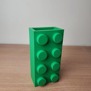K&ouml;nnte beinhalten: Ein leuchtend gr&uuml;ner Stifthalter, der einem gro&szlig;en Baustein nachempfunden ist. Der Halter hat acht kreisf&ouml;rmige Noppen auf der Vorderseite und besteht aus massivem gr&uuml;nem Kunststoff. Das Objekt steht auf einer Holzoberfl&auml;che.