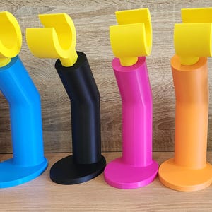 May include: Six 3D printed phone stands in different colors: red, blue, black, pink, orange, and green. Each stand has a yellow hook at the top.