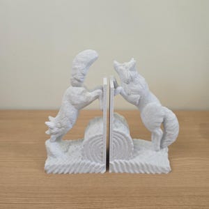 Op de afbeelding: Een paar witte vossen boekensteunen. Elke boekensteun toont een vos die op zijn achterpoten staat, aan weerszijden van een logvormige scheidingswand. De boekensteunen zijn gemaakt van een getextureerd, steenachtig materiaal.