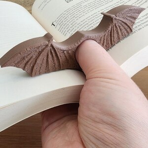 May include: A brown bat-shaped bookmark is inserted into a book. The bookmark has detailed wing features. The open book displays text on its pages. The bookmark is held in place by a hand, suggesting its use for marking a page.
