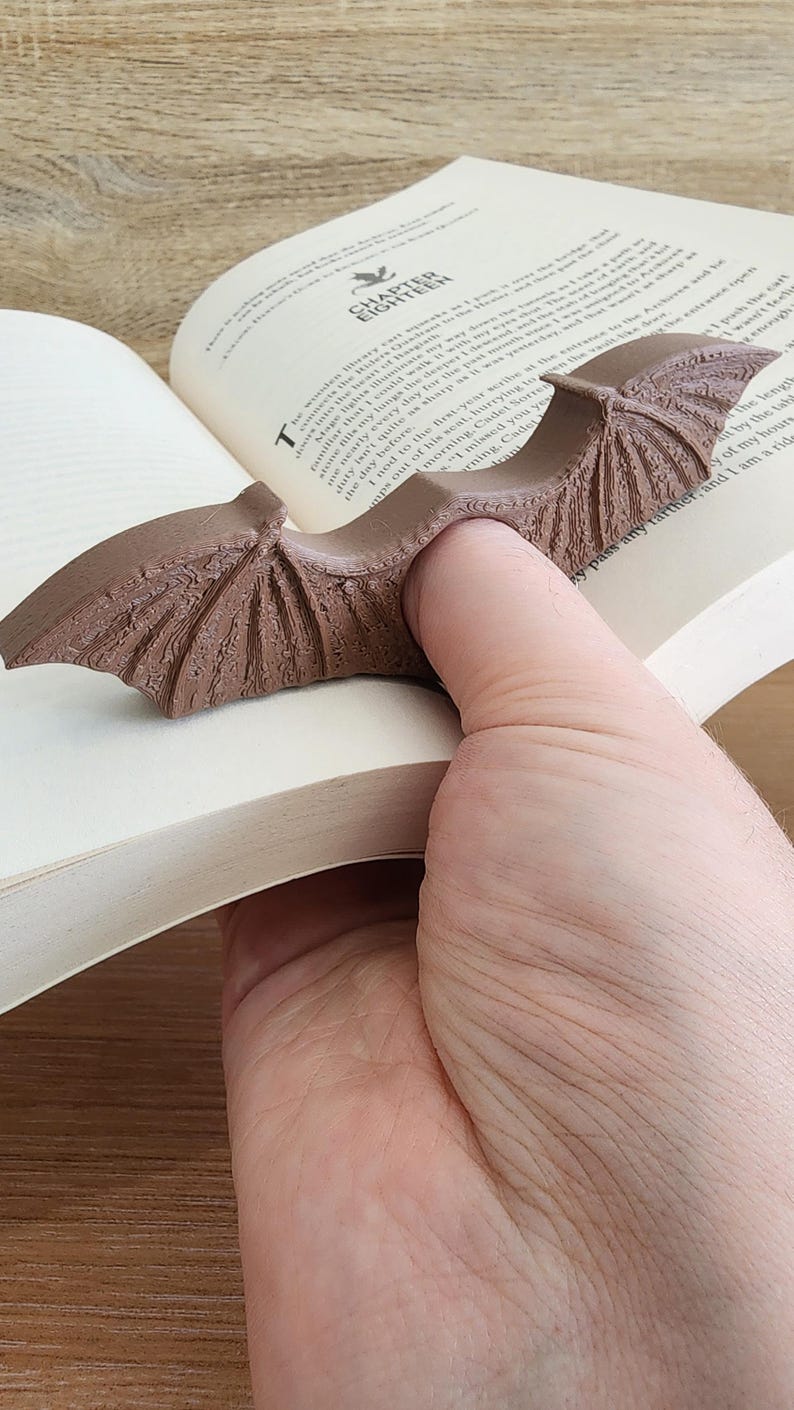 May include: A brown bat-shaped bookmark is inserted into a book. The bookmark has detailed wing features. The open book displays text on its pages. The bookmark is held in place by a hand, suggesting its use for marking a page.