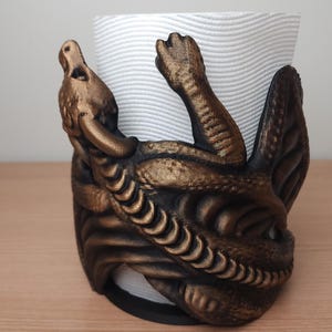 May include: A gold-colored metal paper towel holder shaped like a dragon. The dragon is curled around the paper towel roll and has a textured surface.