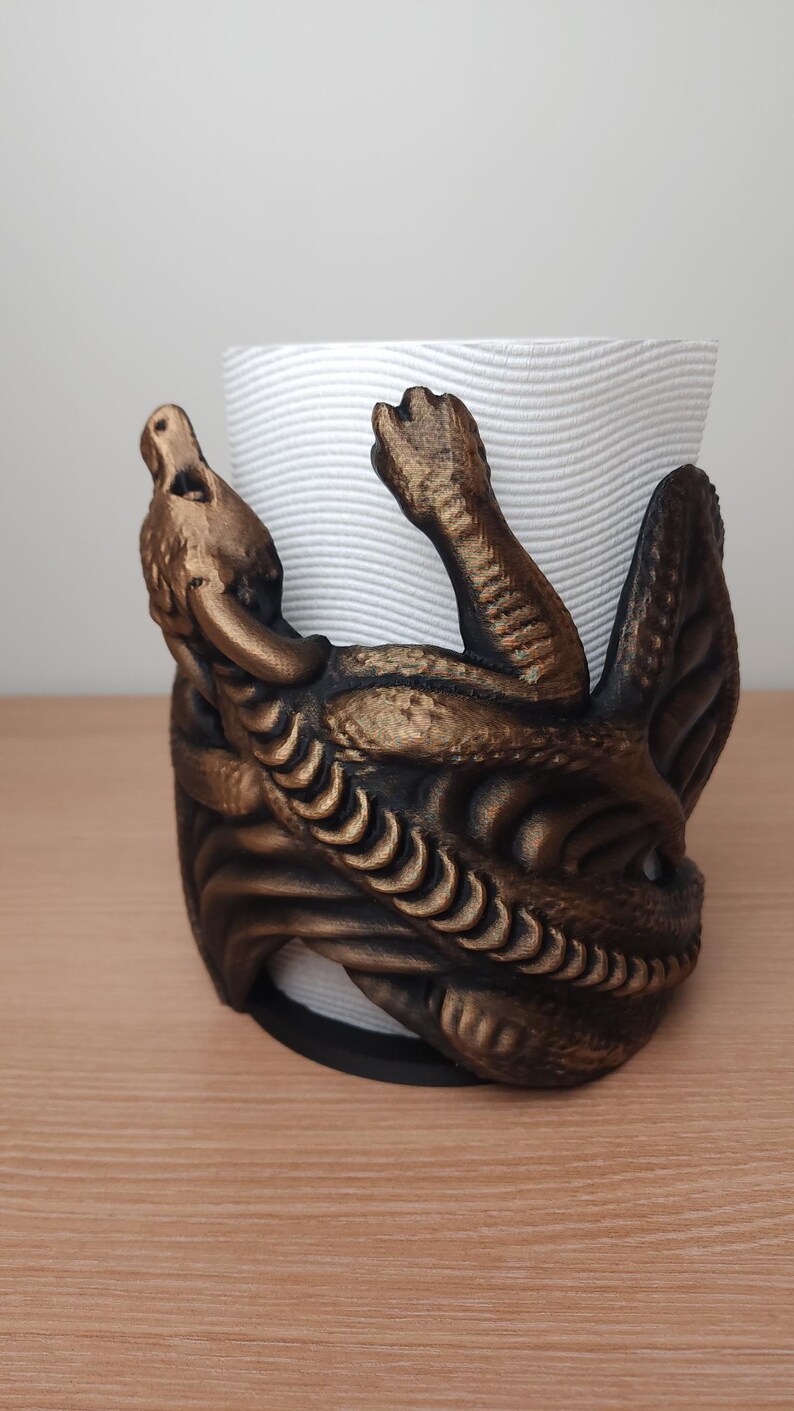 May include: A gold-colored metal paper towel holder shaped like a dragon. The dragon is curled around the paper towel roll and has a textured surface.