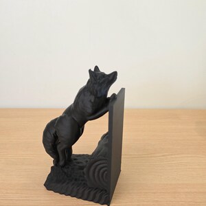 Op de afbeelding: Een zwarte decoratieve boekensteun met een wolf die op een rechthoekige steun klimt. De wolf is gebeeldhouwd met gedetailleerde vacht en staat op een getextureerde basis. De boekensteun is ontworpen om boeken rechtop te houden.