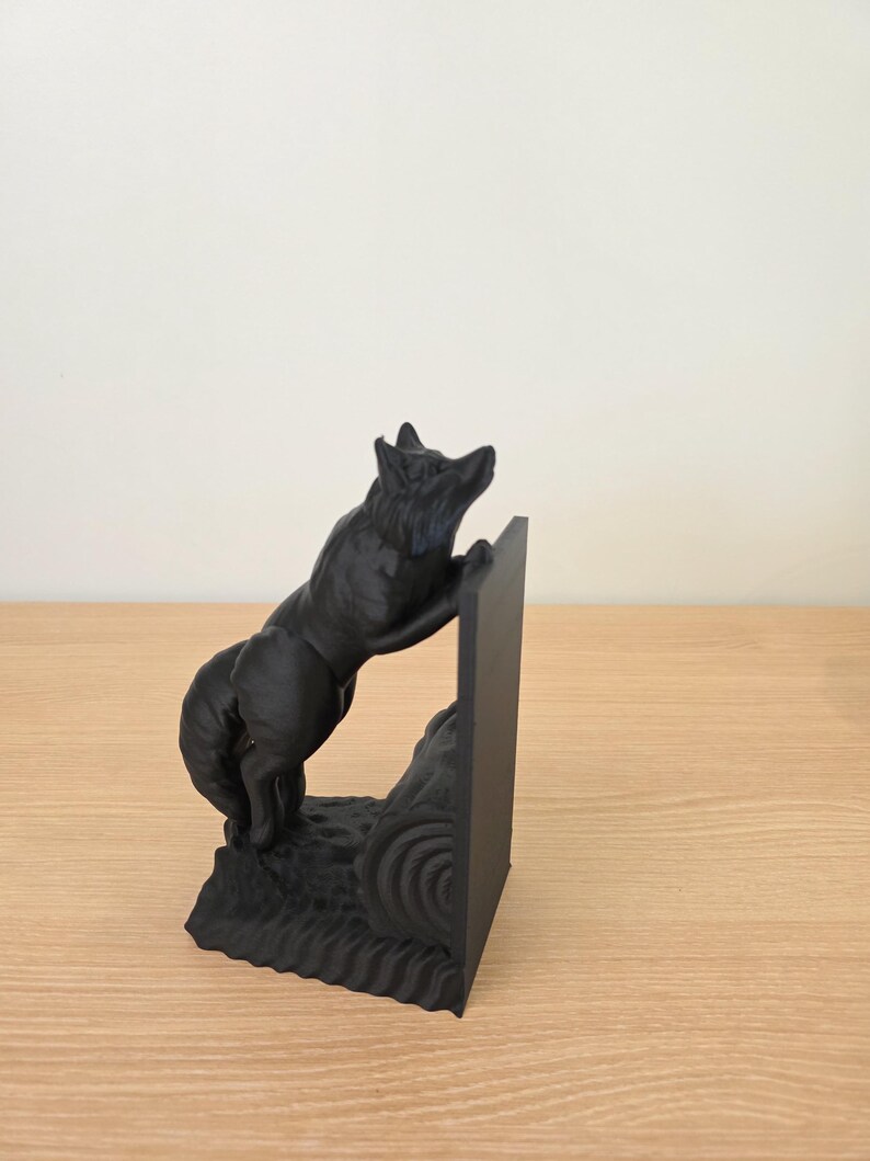 Op de afbeelding: Een zwarte decoratieve boekensteun met een wolf die op een rechthoekige steun klimt. De wolf is gebeeldhouwd met gedetailleerde vacht en staat op een getextureerde basis. De boekensteun is ontworpen om boeken rechtop te houden.