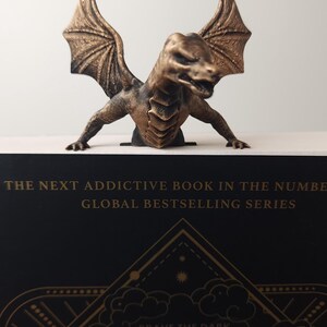 May include: A bronze-colored dragon figurine with outstretched wings sits atop a book. The book has a black cover with gold lettering that reads "ONYX" and "BRAVE THE DARK". The text "THE NEXT ADDICTIVE BOOK IN THE NUMBER GLOBAL BESTSELLING SERIES" is also visible.