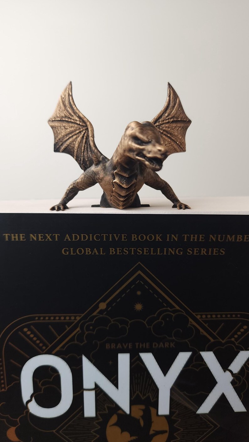 May include: A bronze-colored dragon figurine with outstretched wings sits atop a book. The book has a black cover with gold lettering that reads "ONYX" and "BRAVE THE DARK". The text "THE NEXT ADDICTIVE BOOK IN THE NUMBER GLOBAL BESTSELLING SERIES" is also visible.