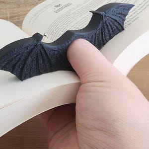 May include: A black bat-shaped bookmark with a textured surface. The bookmark is holding a page open in a book.