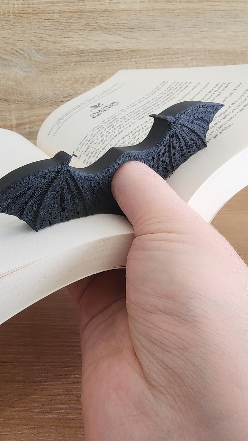 May include: A black bat-shaped bookmark with a textured surface. The bookmark is holding a page open in a book.