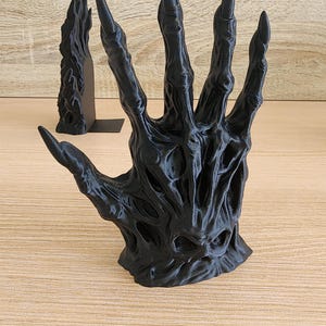 Peut inclure: Sculpture d'une main noire avec de longs doigts pointus. La main est finement d&eacute;taill&eacute;e avec une surface textur&eacute;e et une apparence squelettique. Une deuxi&egrave;me sculpture plus petite est visible en arri&egrave;re-plan.