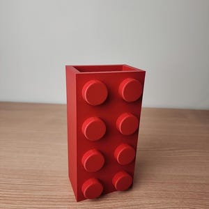 K&ouml;nnte beinhalten: Ein roter, rechteckiger, blockf&ouml;rmiger Beh&auml;lter mit acht kreisf&ouml;rmigen Noppen auf der Vorderseite. Der Beh&auml;lter besteht aus einem einfarbigen roten Material und hat einen hohlen Innenraum. Das Design erinnert an ein klassisches Bausteinspielzeug.