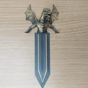 May include: A bronze-colored dragon bookmark with outstretched wings and a detailed body. The dragon sits atop a dark blue bookmark with a pointed bottom. The bookmark is a fantasy-themed item.