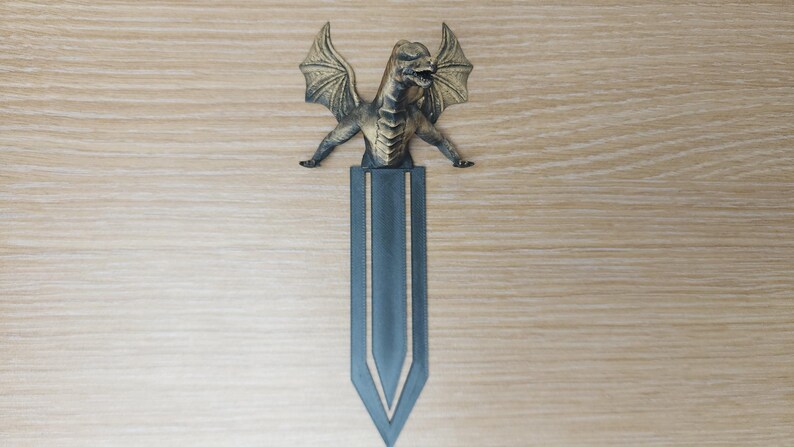 May include: A bronze-colored dragon bookmark with outstretched wings and a detailed body. The dragon sits atop a dark blue bookmark with a pointed bottom. The bookmark is a fantasy-themed item.