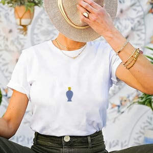 Peut inclure: T-shirt blanc avec un petit motif de flacon de parfum bleu et un bouchon jaune. La personne porte un chapeau de paille, des colliers et des bracelets dor&eacute;s. Le fond pr&eacute;sente un mur &agrave; motifs floraux et des plantes en pot.