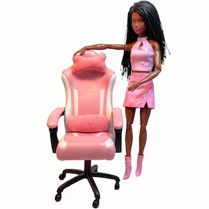 Könnte beinhalten: Ein rosa Gaming-Stuhl mit weißen Akzenten und einem rosa Kissen. Der Stuhl hat eine Kopfstütze und ein Lendenkissen. Eine Puppe steht neben dem Stuhl.