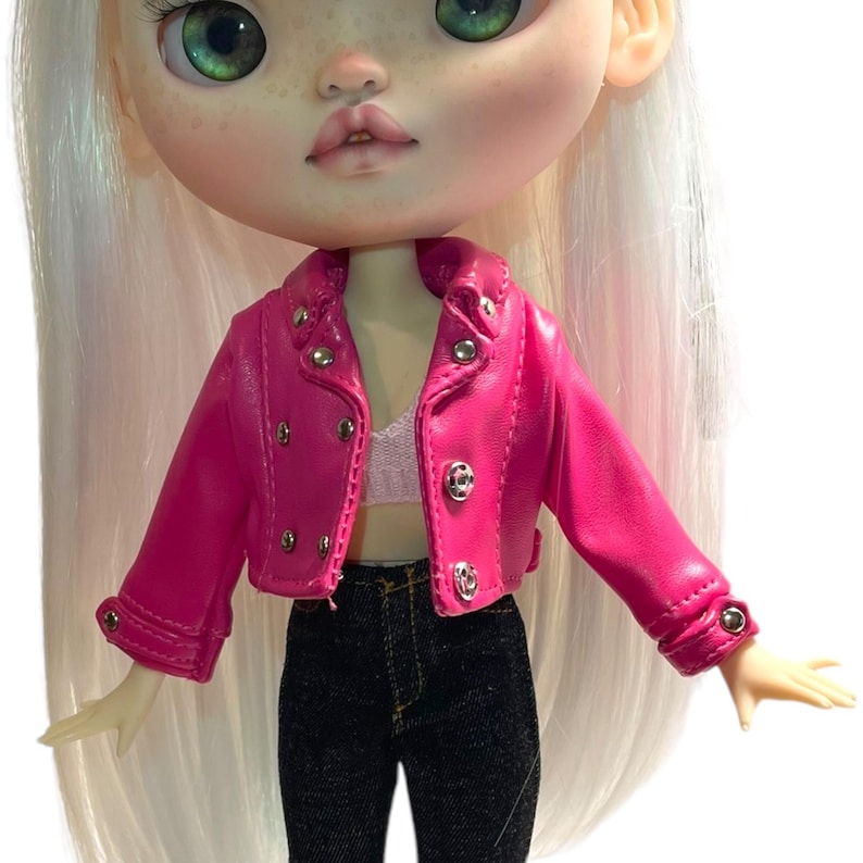 Pu&ograve; includere: Una bambola con una giacca rosa acceso in similpelle con bottoni argentati, un top bianco e pantaloni di jeans neri. La bambola ha lunghi capelli bianchi, occhi verdi e carnagione chiara. La giacca &egrave; in classico stile biker.