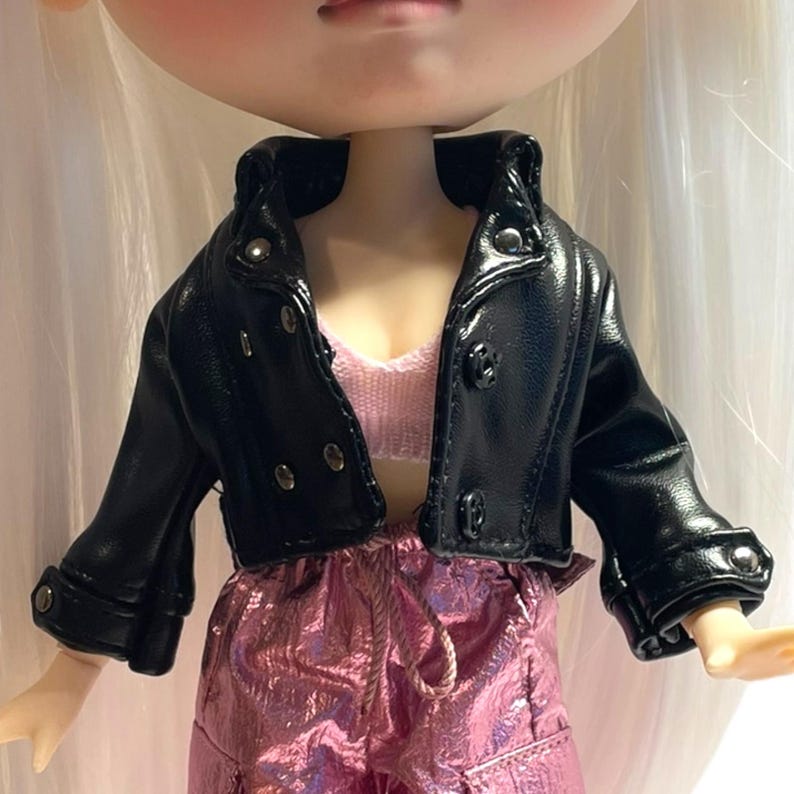 Pu&ograve; includere: Una bambola indossa una giacca in similpelle nera con bottoni argentati e un top rosa. La bambola indossa anche pantaloni metallizzati rosa. La giacca ha un classico stile biker con colletto a revers e polsini abbottonati.