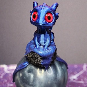 Peut inclure: Figurine de dragon bleu avec de grands yeux rouges. Le dragon est perché sur une base sphérique bleu foncé. La figurine a un aspect texturé, imprimé en 3D. La tête du dragon est droite, avec de petites cornes et une expression amicale.