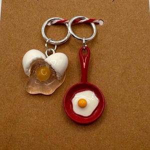 May include: A pair of novelty earrings featuring a fried egg and a fried egg in a red pan. The earrings are attached to a brown card with a butterfly cutout. The earrings are attached to a silver hoop.