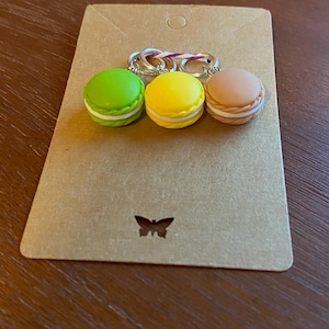 May include: Three macaron-shaped stitch markers in green, yellow, and brown. The markers are on a brown card with a butterfly logo.