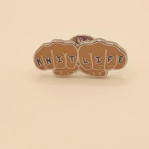 May include: A silver pin with two fists, each with a knuckle tattoo. The left fist says "KNIT" and the right fist says "LIFE".