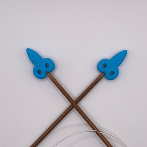 May include: A pair of blue and brown circular knitting needles with a cable. The needles have a blue plastic tip with a decorative design.