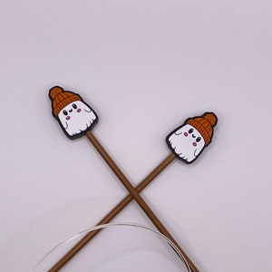 May include: Two brown knitting needles with gold tips are crossed, featuring white ghost-shaped toppers wearing orange knit caps. A clear, flexible cable connects the needles, creating a circular knitting tool. The background is plain white.