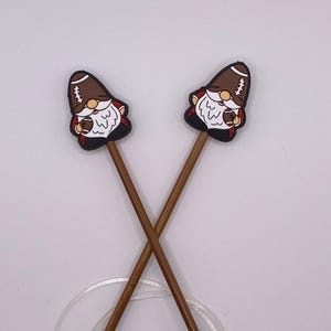 May include: A pair of brown bamboo knitting needles with gold tips and football gnome-shaped end caps. The needles are crossed and a clear cord is wrapped around them.