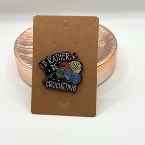 May include: A pin featuring the text "I'd rather be crocheting" with colorful yarn balls. The pin is attached to a brown card with a butterfly graphic. The pin is set on a copper-colored, circular surface.