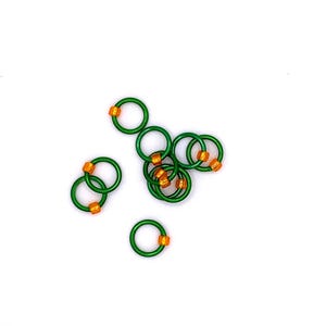 May include: A group of green and orange stitch markers, also known as knitting rings, are scattered on a white surface.