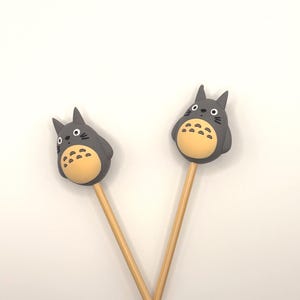 May include: Two gray Totoro-shaped toppers with yellow bellies and black eyes are attached to wooden sticks. The toppers are positioned against a white background. The sticks are angled, creating an X shape.