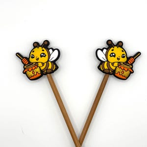 May include: Two decorative bee-themed toppers. Each topper features a cartoon bee with yellow, black, and white accents, holding a honey pot. The toppers are attached to brown wooden sticks, perfect for cupcakes or other treats.