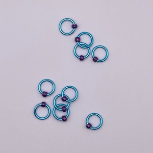 May include: A set of eight teal-colored metal rings with purple beads. The rings are used for knitting or crocheting.