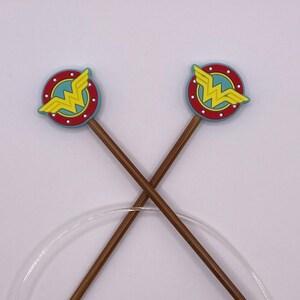 May include: Two brown knitting needles with gold tips are crossed over each other. Each needle has a Wonder Woman logo at the top, featuring a yellow lightning bolt on a blue background, surrounded by a red circle with white stars.