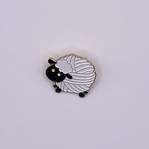 May include: Gold-tone enamel pin of a white sheep with black face and yellow eyes, shaped like a ball of yarn.