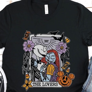 Pu&ograve; includere: Maglietta nera con una grafica di Jack Skellington e Sally di The Nightmare Before Christmas, raffigurati come la carta dei Tarocchi degli Amanti. La carta ha uno sfondo viola con un sole e una luna, e il testo "THE LOVERS" &egrave; stampato sotto l'immagine.