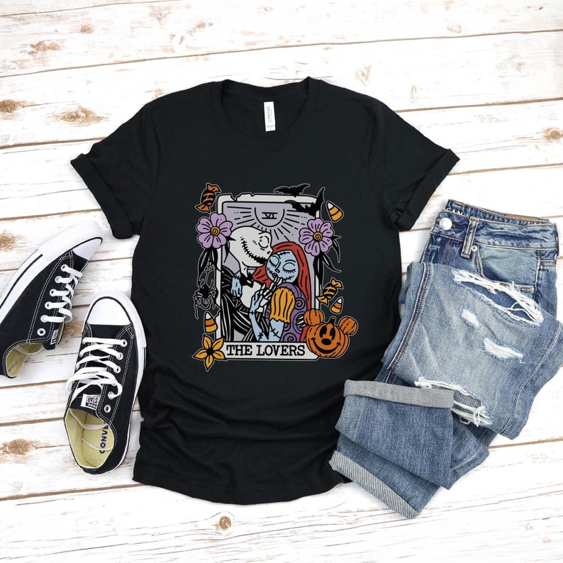 Pu&ograve; includere: Maglietta nera con una grafica di Jack Skellington e Sally di The Nightmare Before Christmas, raffigurati come la carta dei Tarocchi degli Amanti. La carta ha uno sfondo viola con un sole e una luna, e il testo "THE LOVERS" &egrave; stampato sotto l'immagine.