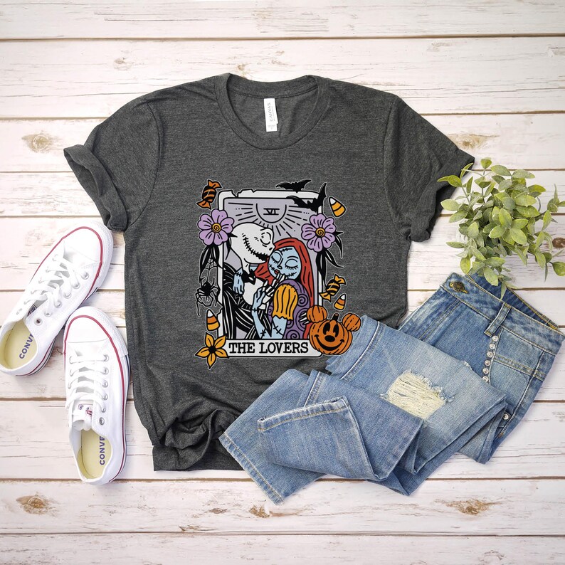 Pu&ograve; includere: Una maglietta grigio scuro con una grafica di Jack Skellington e Sally di The Nightmare Before Christmas. La grafica &egrave; una carta dei tarocchi con il testo "The Lovers" e il numero "VI" su di essa. La carta &egrave; circondata da immagini a tema Halloween, tra cui mais dolce, pipistrelli e zucche.