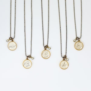 May include: Five gold necklaces with small round pendants featuring handwritten letters A, B, C, D, and E. Each pendant is on a delicate chain with a small pearl accent.