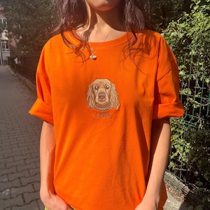 May include: Bright orange t-shirt featuring an embroidered dog portrait with the name "LENA" below. The shirt has short, rolled-up sleeves and a crew neck. The dog's fur is a mix of brown and tan.